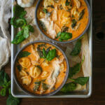 Two bowls of chicken tortellini soup on a metal tray with baby kale and basil scattered around.