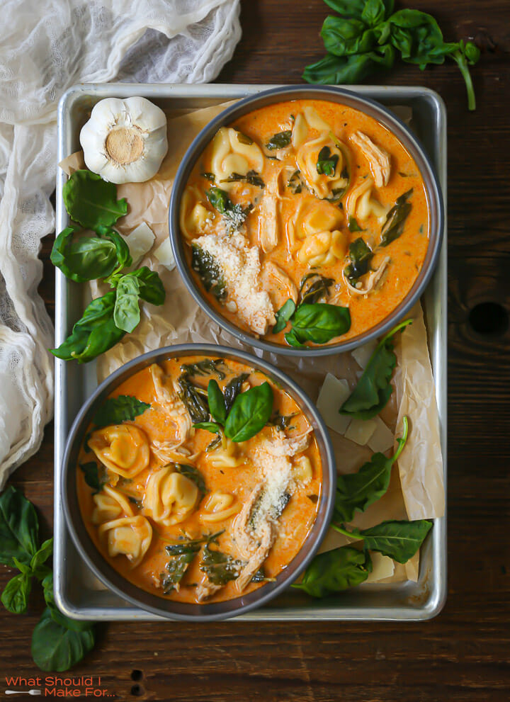 Two bowls of chicken tortellini soup on a metal tray with baby kale and basil scattered around.