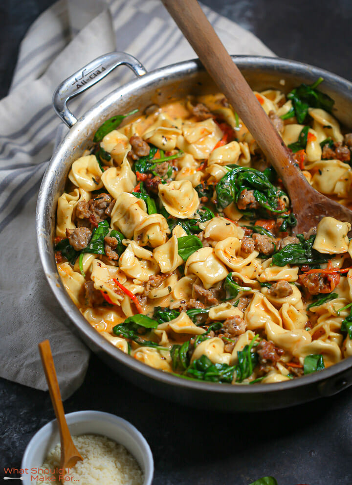 A pan of Creamy Sausage Tortellini with Spinach and Tomatoes stirred with a wooden spoon.