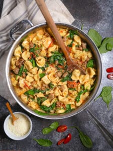 Overhead shot of One Pot Creamy Sausage Tortellini with Spinach and Tomatoes with a wooden spoon stirring the pot.