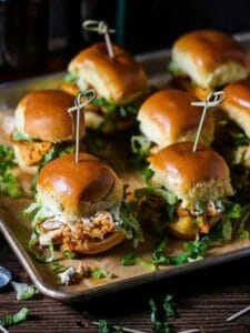 Buffalo Chicken Sliders skewered with knotted toothpicks on a sheet tray with beer in the background and a bottle cap on the table.
