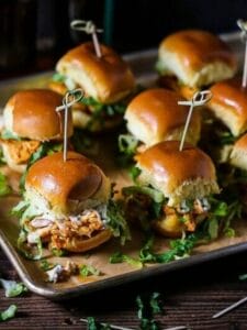 Buffalo Chicken Sliders skewered with knotted toothpicks on a sheet tray with beer in the background and a bottle cap on the table.