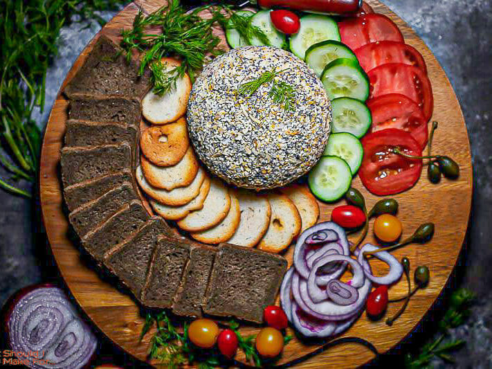 Overhead shot of Everything Bagel Smoked Salmon and Cream Cheese Ball on a platter with veggies, pumpernickel toasts and herbs.