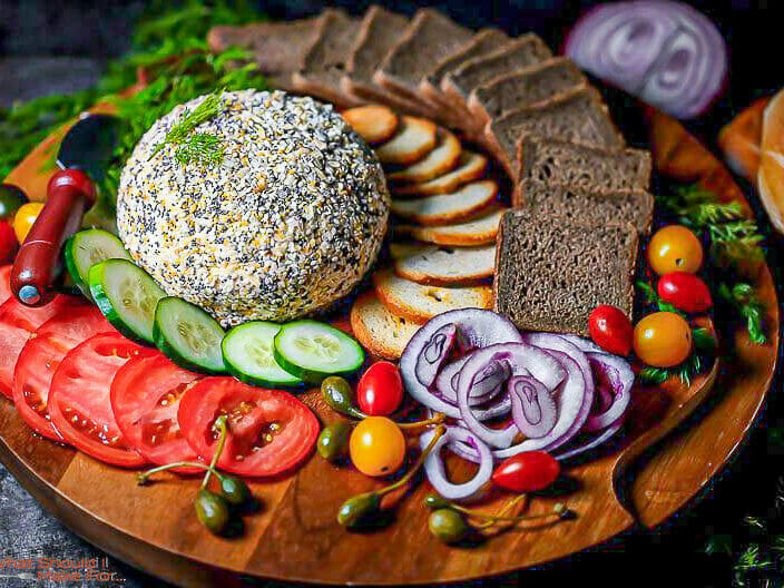 Close up of Everything Bagel Smoked Salmon and Cream Cheese Ball on a platter with veggies, pumpernickel toasts and herbs.